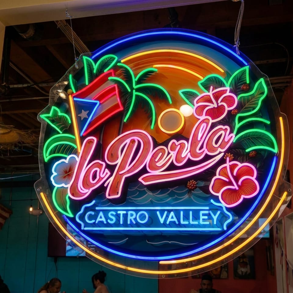 Colorful neon La Perla Castro Valley sign featuring tropical flowers, palm leaves, a sunset, and Puerto Rican flag inside a restaurant.