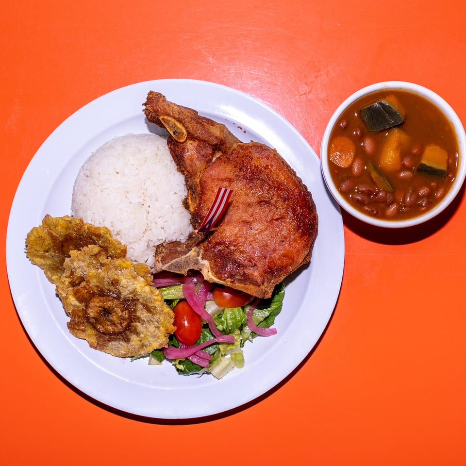 Plate of fried pork, white rice, tostones, and salad with pickled onions, served alongside a bowl of bean and vegetable stew.