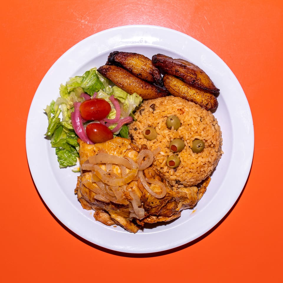 Plate of seasoned rice with olives, grilled chicken, fried plantains, and lettuce salad on an orange table.