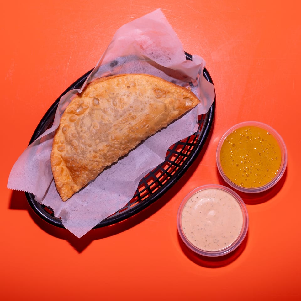Crispy folded empanada in a lined basket with yellow and creamy dipping sauces on an orange table.