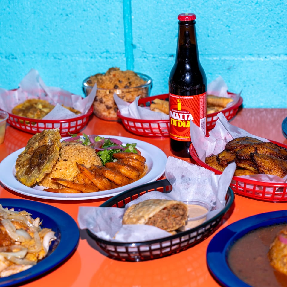 Colorful restaurant spread featuring rice, grilled meats, fried plantains, side dishes, and a bottle of Malta India on a bright table.