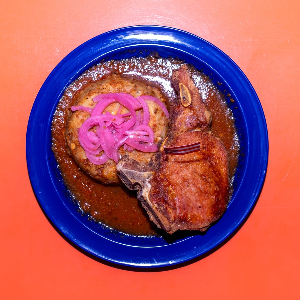 Blue plate of grilled meat and a patty in brown sauce, topped with pickled red onions, on an orange table.
